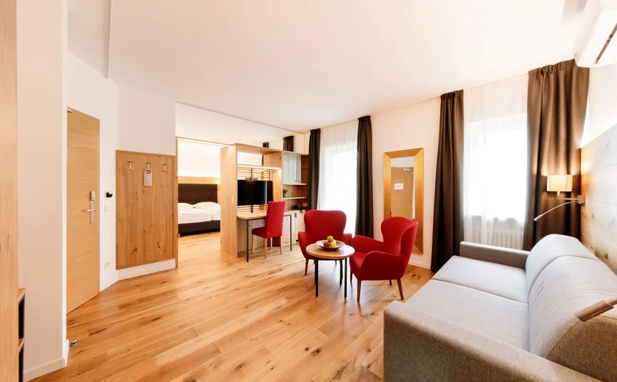 Bright hotel room with wooden floor, red armchairs, and gray sofa