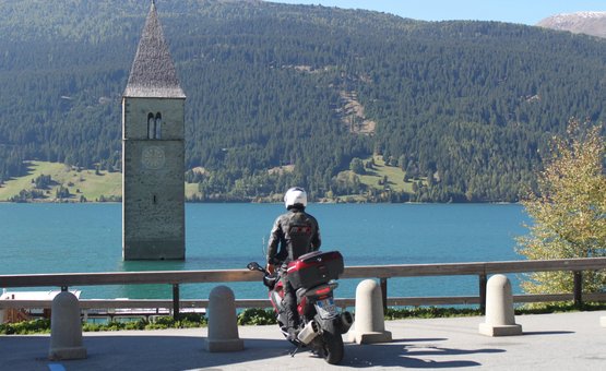 Motorradfahrer blickt auf Turm im See vor bewaldeten Bergen