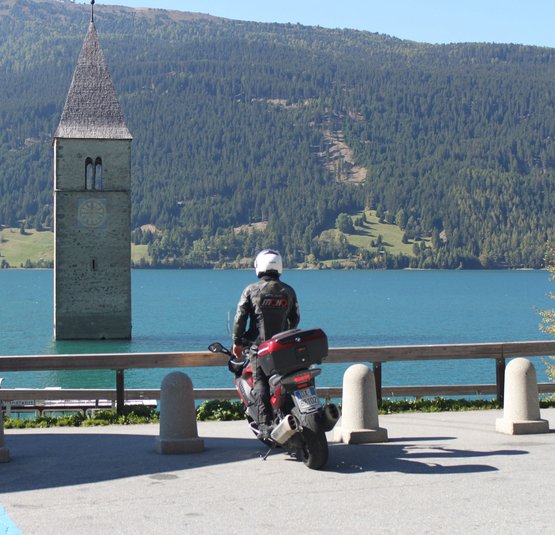 Motorradfahrer blickt auf Turm im See vor bewaldeten Bergen