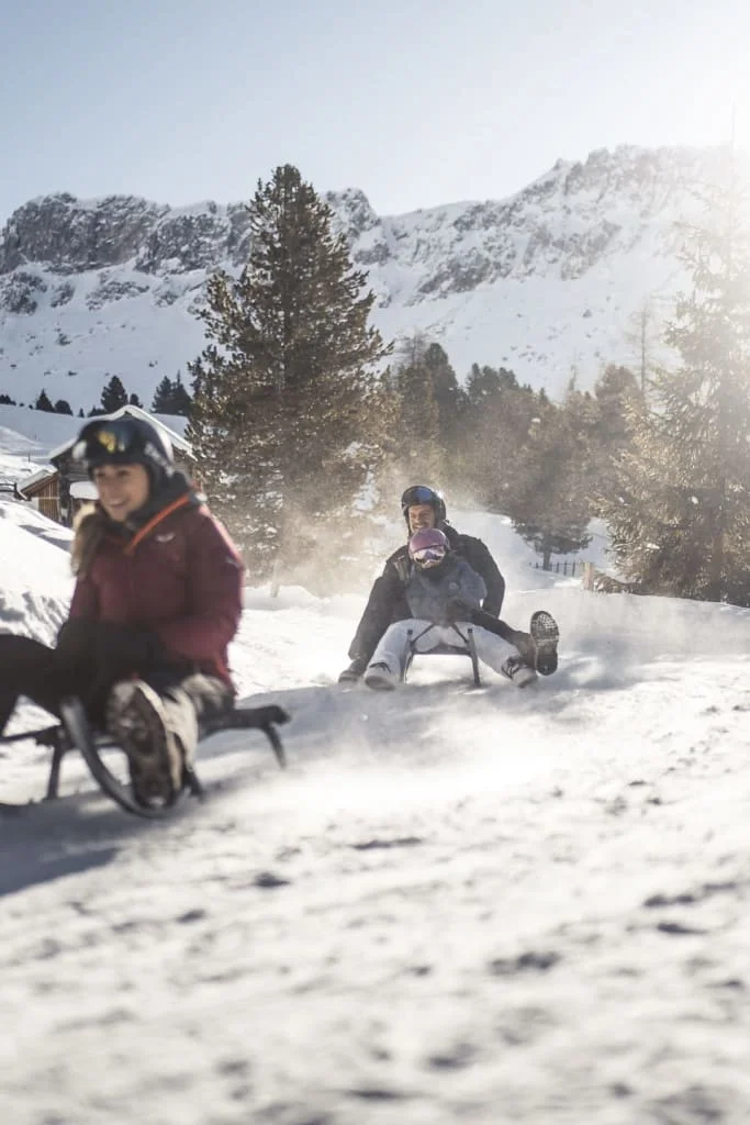 7=6 Jonathan´s Winter Wohlfühlwochen Menschen rodeln bei sonnigem Wetter auf schneebedecktem Hang mit Bergen im Hintergrund