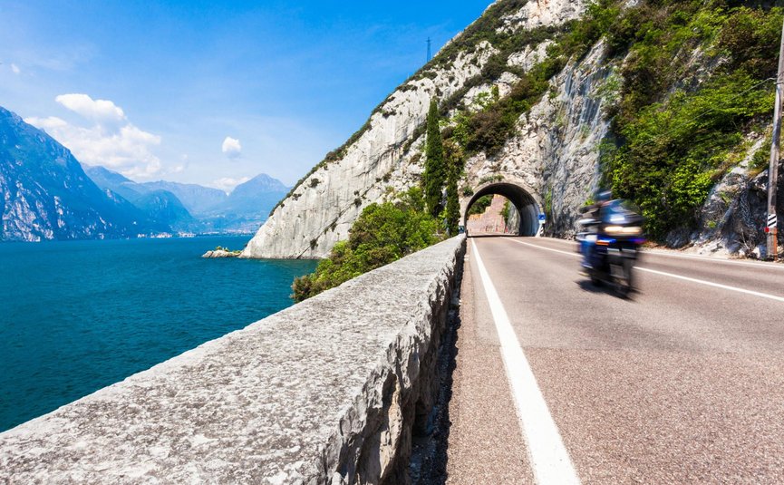 Motorradurlaub in Val di Sole – Brenta Dolomiten – Gardasee © TonelliHotels Motorradfahrer fährt auf Straße neben See und Bergtunnel bei klarem Himmel