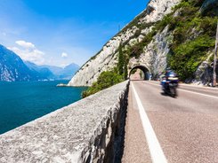 Val di Sole – Brenta Dolomiten – Gardasee © TonelliHotels Motorradfahrer fährt auf Straße neben See und Bergtunnel bei klarem Himmel