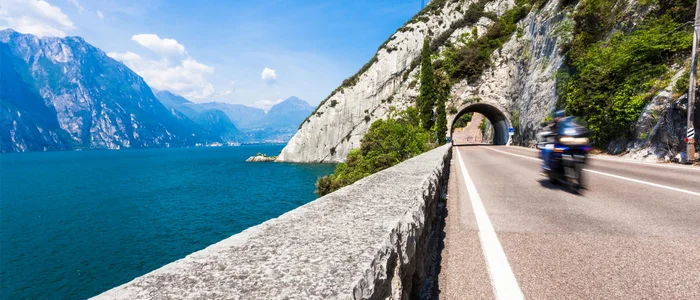Motorradfahrer fährt auf Straße neben See und Bergtunnel bei klarem Himmel