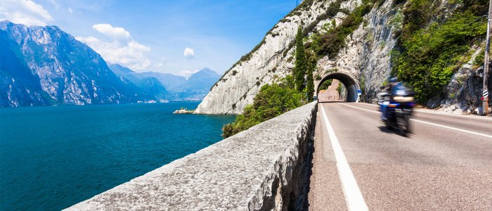 Motorcyclist riding on road beside lake and mountain tunnel under clear sky