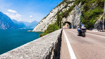 Val di Sole – Brenta Dolomiten – Gardasee © TonelliHotels Motorradfahrer fährt auf Straße neben See und Bergtunnel bei klarem Himmel