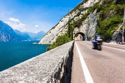 Motorradfahrer fährt auf Straße neben See und Bergtunnel bei klarem Himmel