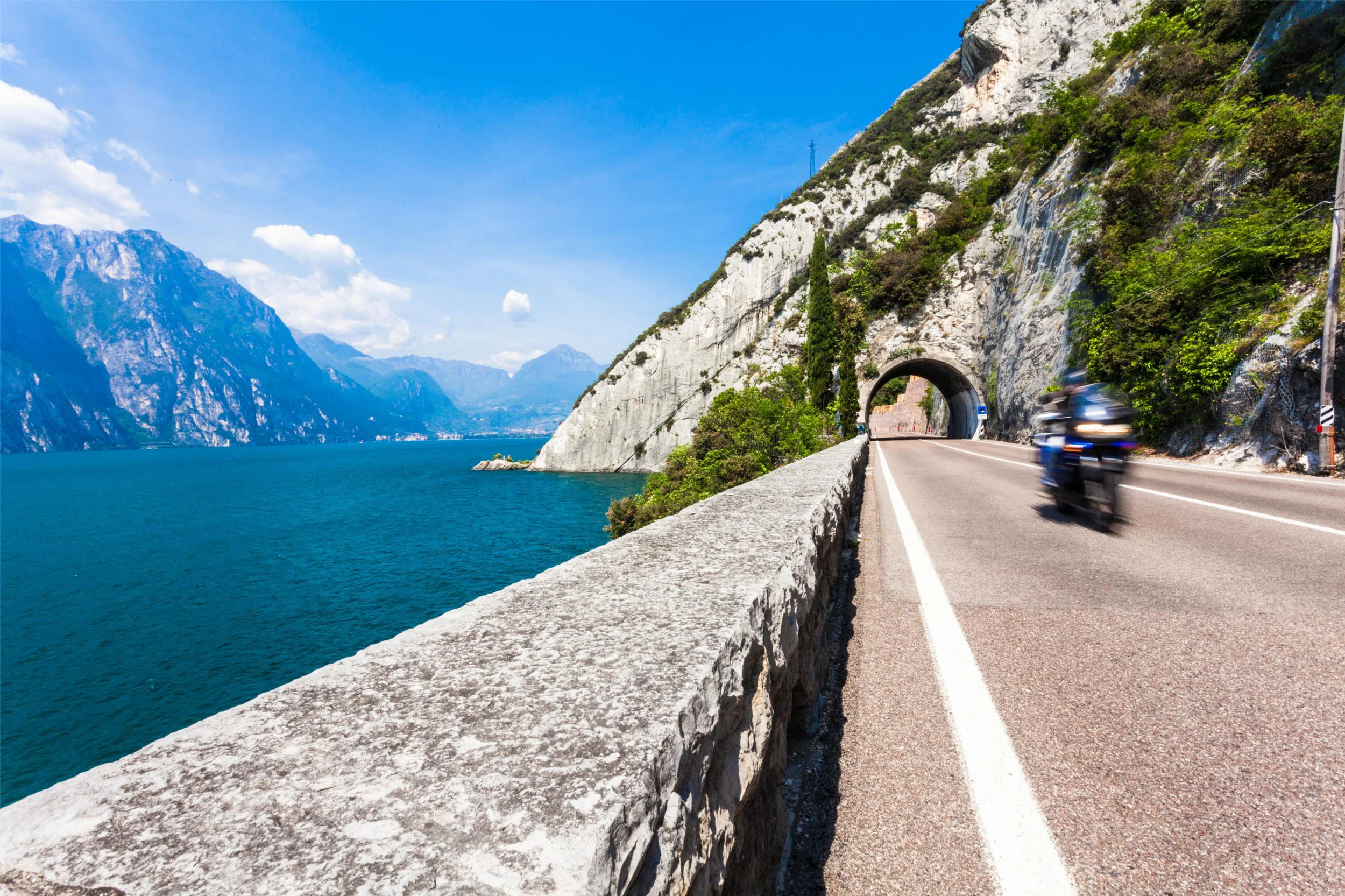 Hotels © TonelliHotels Motorcyclist riding on road beside lake and mountain tunnel under clear sky