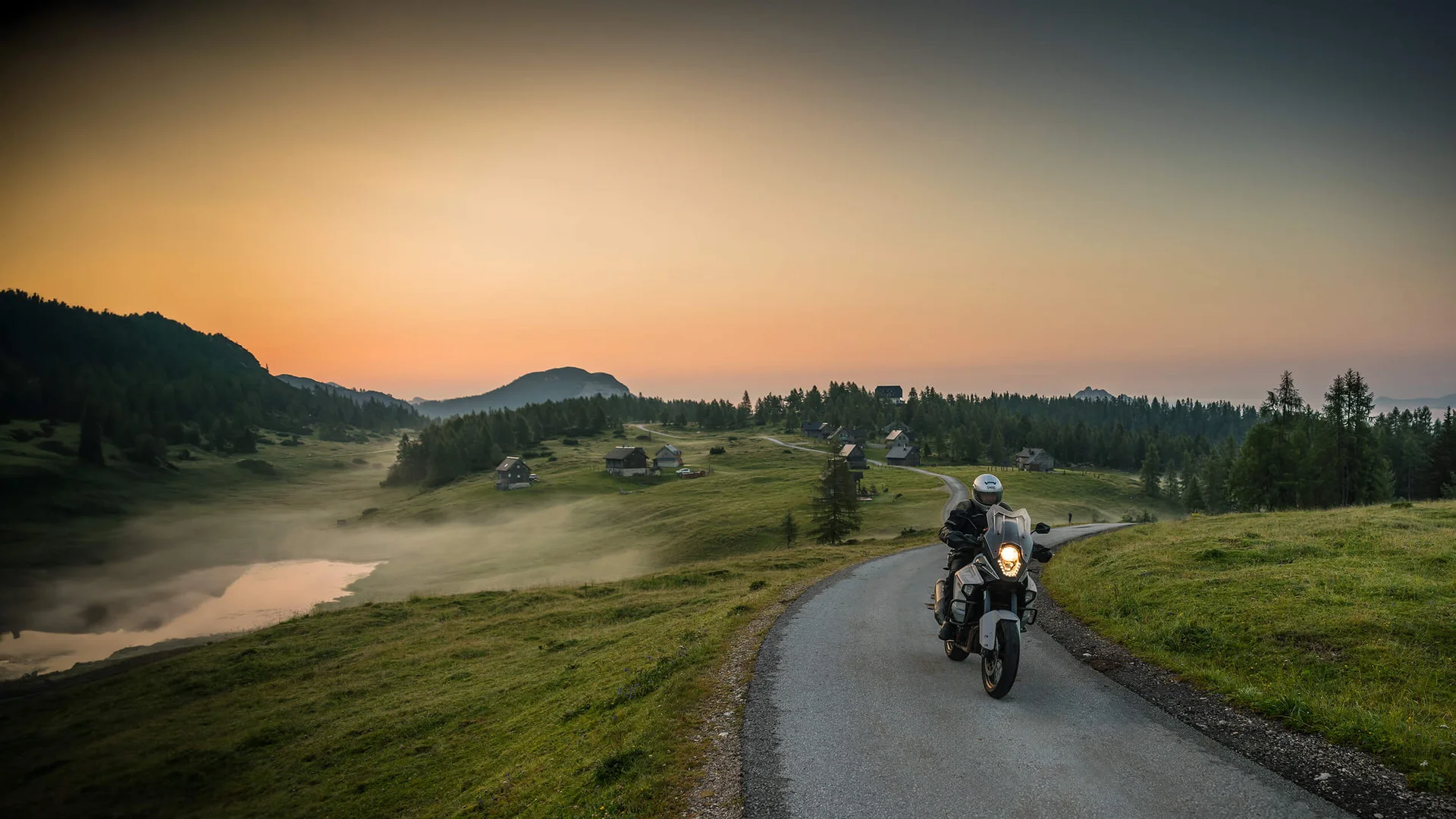 Motorradfahrer fährt auf Landstraße bei Sonnenuntergang mit Hütten und Bergen