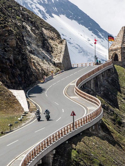 Kurvige Bergstraße mit Motorradfahrern und historischen Grenzsteinen bei Schnee