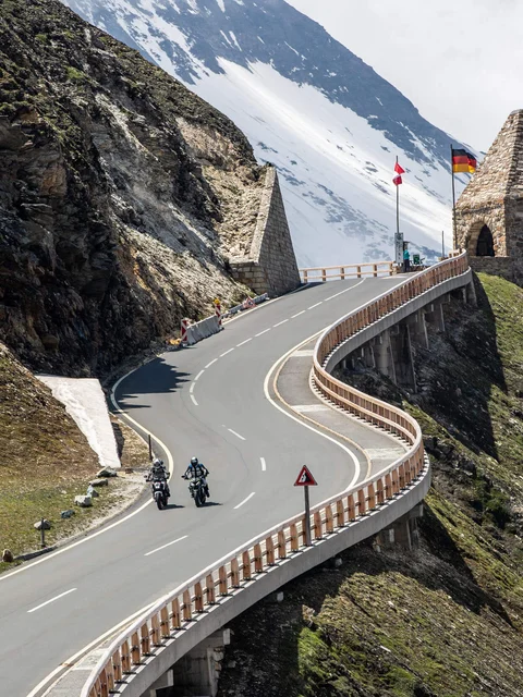 Curvy mountain road with motorcyclists and historic border markers in snowy area