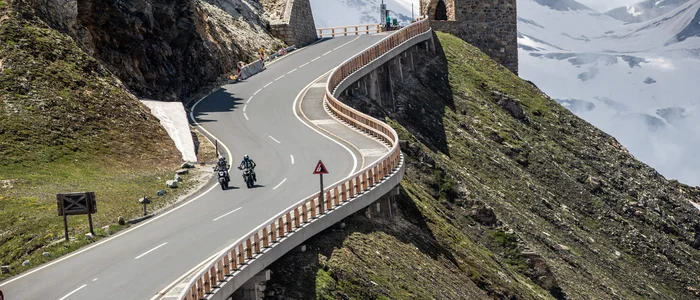 Kurvige Bergstraße mit Motorradfahrern und historischen Grenzsteinen bei Schnee