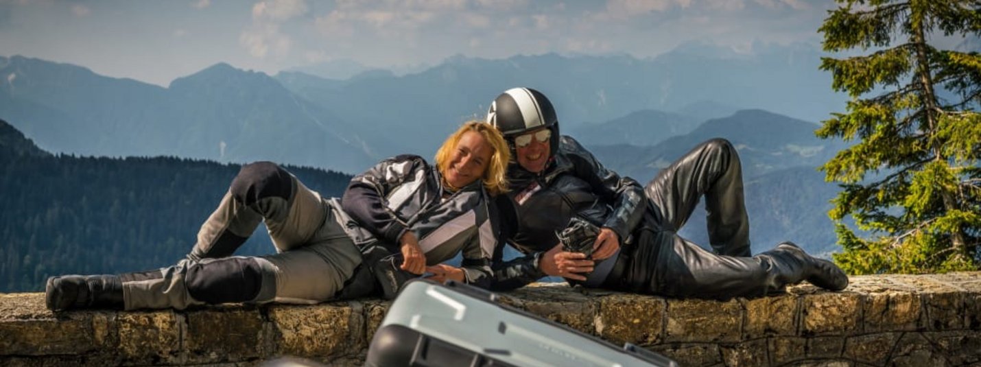 Special-Bike-Week Zwei Motorradfahrer in Schutzkleidung liegen auf einer Steinmauer mit Bergpanorama