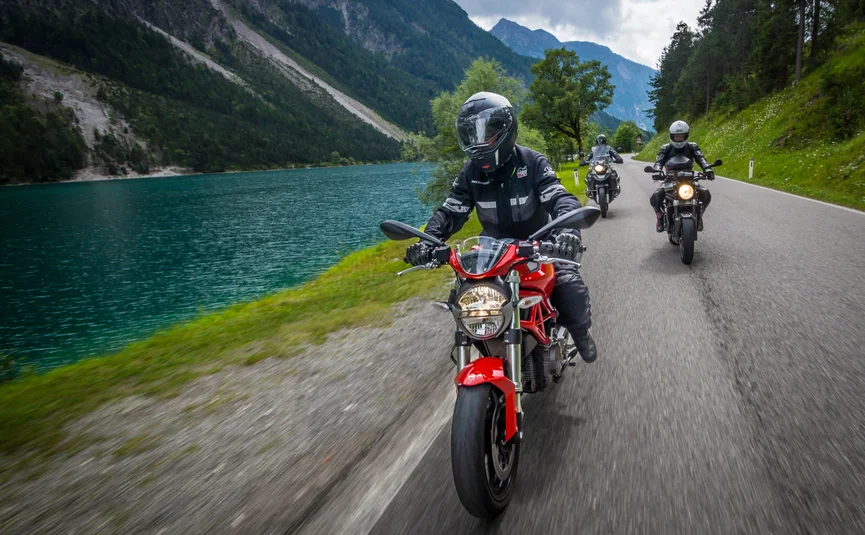 MoHo Schönauer Hof- Tour 7 Kulturtour Drei Motorradfahrer fahren auf einer Straße neben einem See und Bergen.
