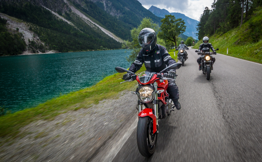 Drei Motorradfahrer fahren auf einer Straße neben einem See und Bergen.