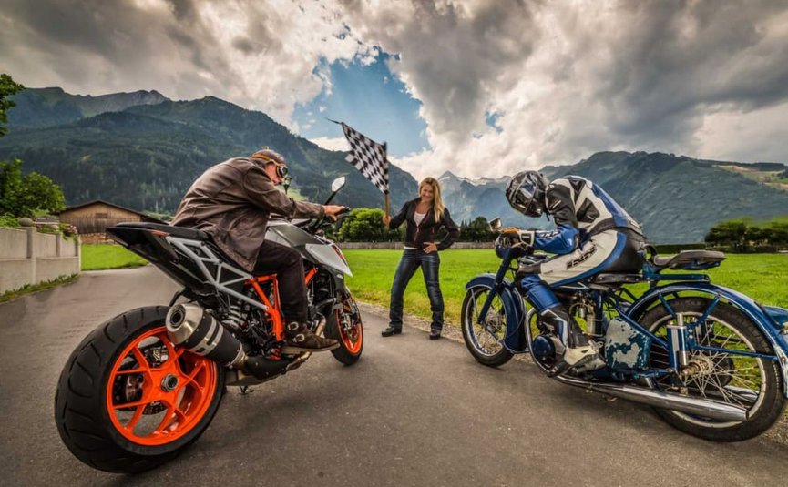 Zwei Motorradfahrer warten auf Startsignal von Frau mit Zielflagge auf Landstraße