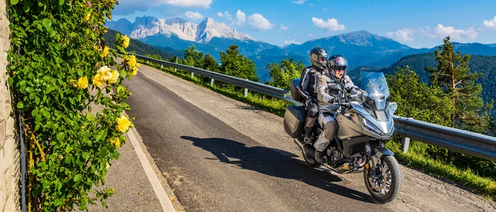 Two motorcyclists riding a mountain road with flowers and mountains in background