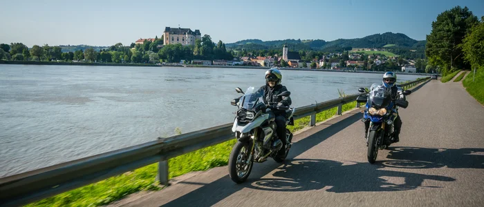 Zwei Motorradfahrer fahren neben einem Fluss mit einer Burg im Hintergrund