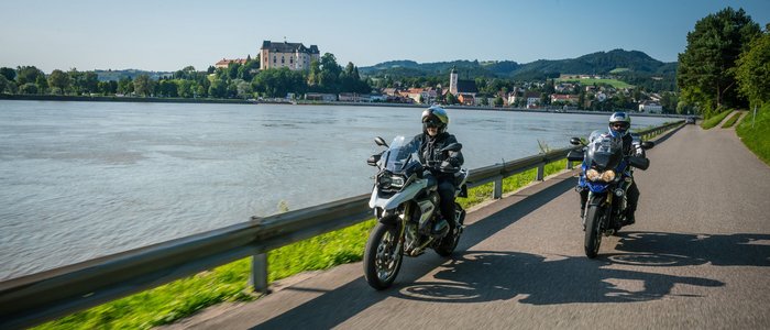 Zwei Motorradfahrer fahren neben einem Fluss mit einer Burg im Hintergrund