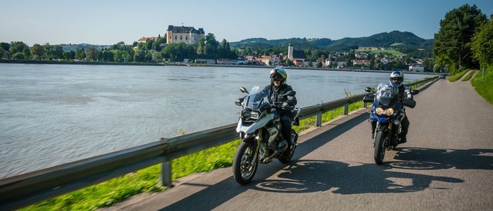 Mühlviertel © Moppetfoto.de Twee motorrijders rijden langs een rivier met een kasteel op de achtergrond