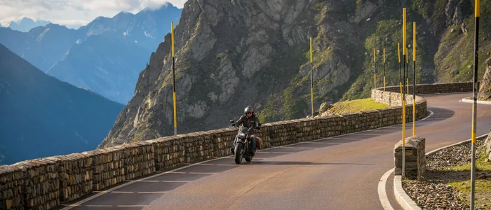 Motorradfahrer auf kurvenreicher Bergstraße mit Steinmauer und Bergen im Hintergrund