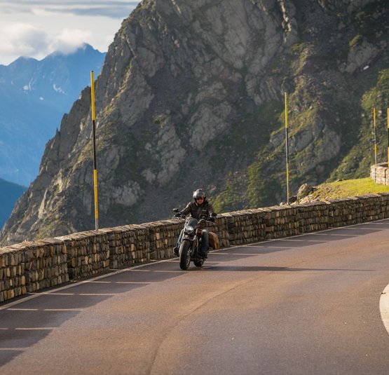 Motorradfahrer auf kurvenreicher Bergstraße mit Steinmauer und Bergen im Hintergrund
