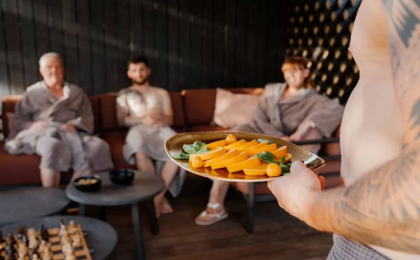 Sweaty Weekends Mann mit Tattoo hält Teller mit Obst vor drei Personen in Bademänteln