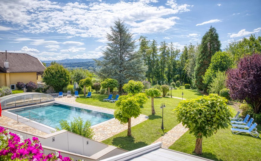 Schwimmbad mit Liegestühlen und gepflegtem Garten mit Bäumen und Bergen im Hintergrund.