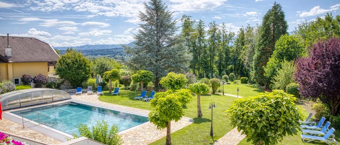 Schwimmbad mit Liegestühlen und gepflegtem Garten mit Bäumen und Bergen im Hintergrund.