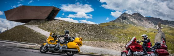 Motorvakantie op de Arlberg - drielandenpunt © Moppetfoto.de Twee motorrijders op bergweg met bergen en modern gebouw op de achtergrond