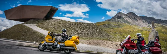 Zwei Motorradfahrer auf Bergstraße mit Bergen und modernem Gebäude im Hintergrund