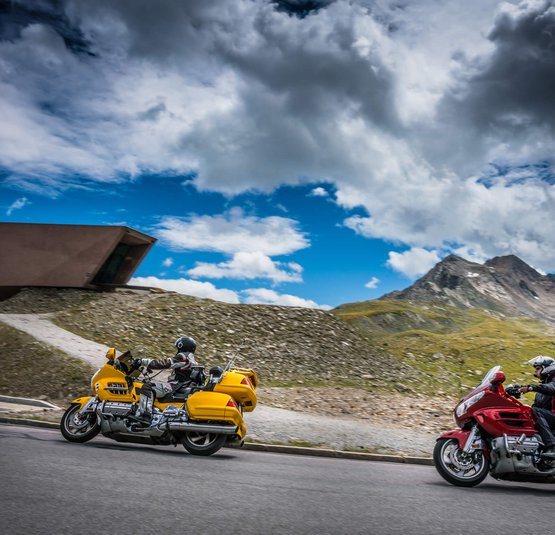 Zwei Motorradfahrer auf Bergstraße mit Bergen und modernem Gebäude im Hintergrund