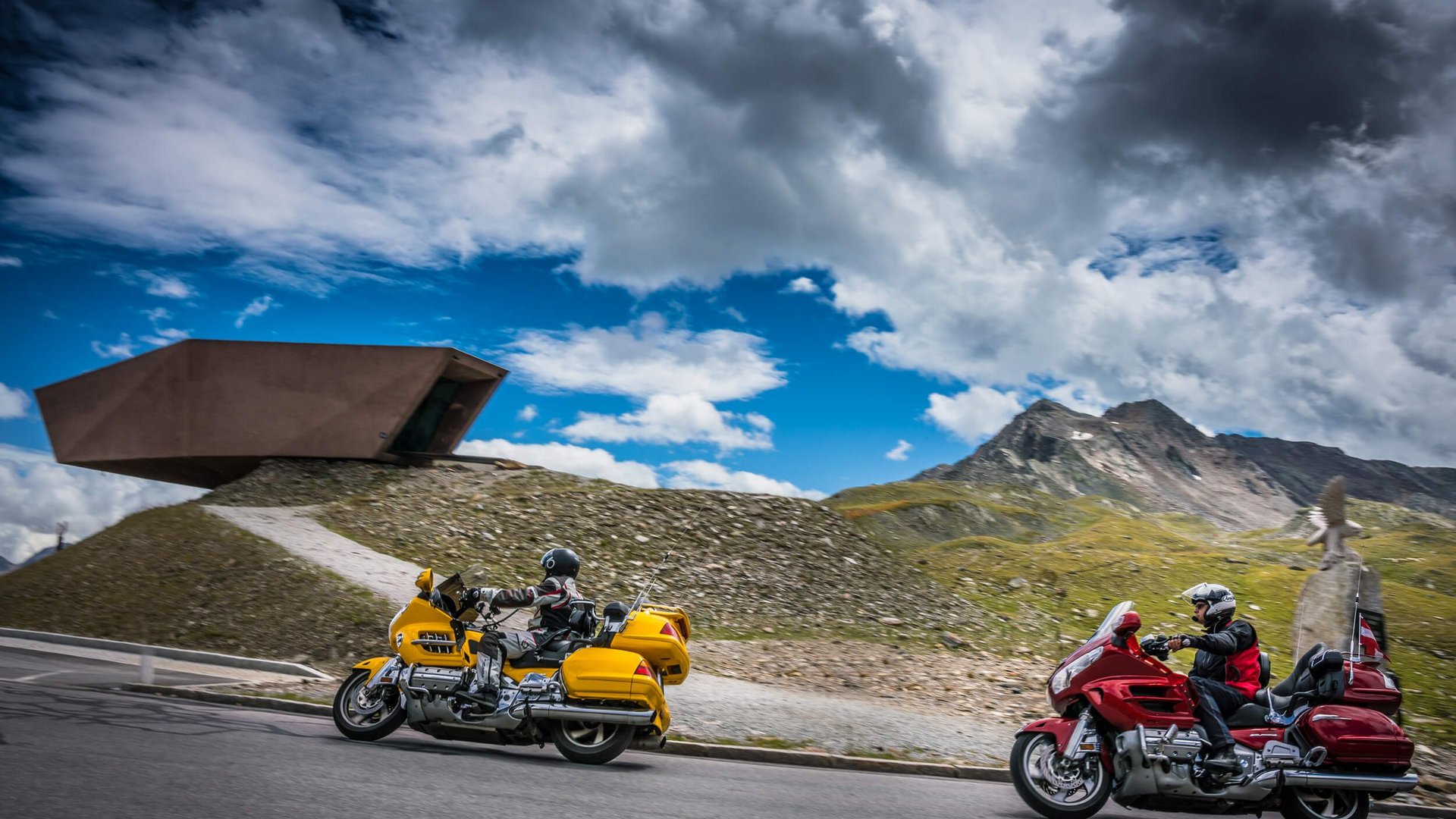 Zwei Motorradfahrer auf Bergstraße mit Bergen und modernem Gebäude im Hintergrund