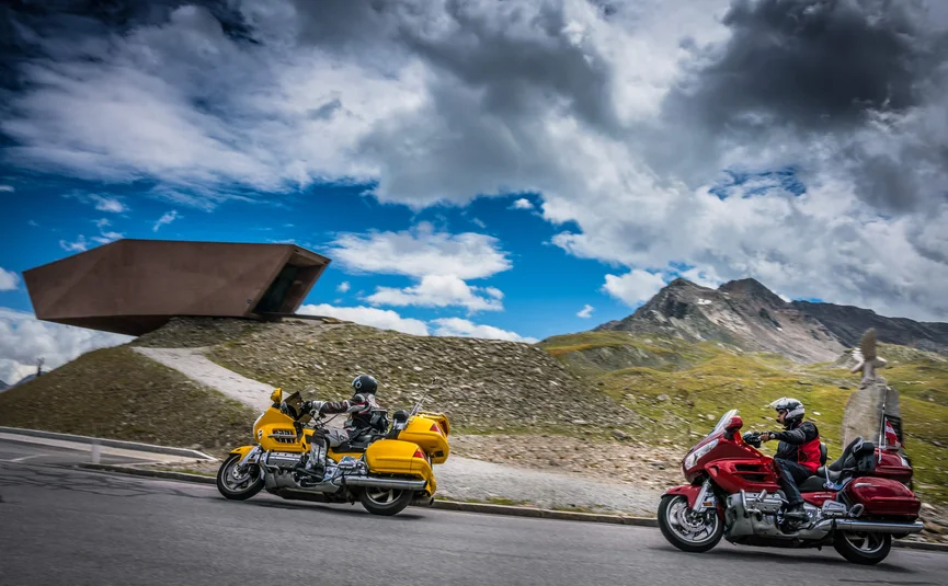 Two motorcyclists riding on mountain road with peaks and modern structure in background