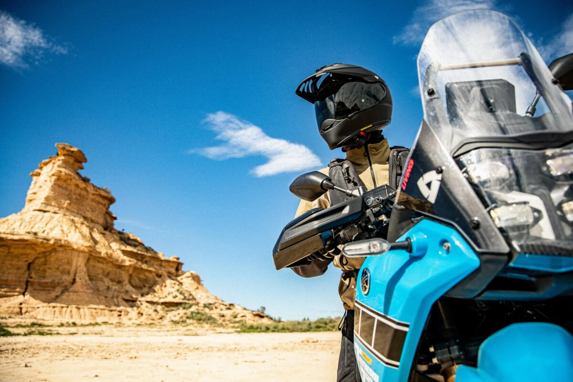 Motorradfahrer mit Helm und Jacke vor felsiger Wüstenlandschaft unter blauem Himmel