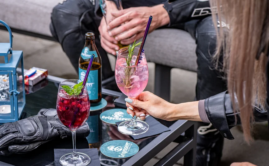 Zwei verschiedene Cocktails und eine Bierflasche auf einem Tisch mit Personen im Hintergrund