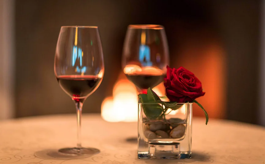 Two wine glasses and a red rose in a glass vase by a fireplace