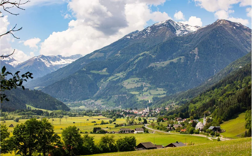 Dorf mit grünen Wiesen und schneebedeckten Bergen unter bewölktem Himmel