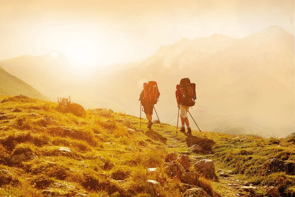 Zwei Wanderer mit Rucksäcken wandern auf einem Bergpfad im Sonnenlicht