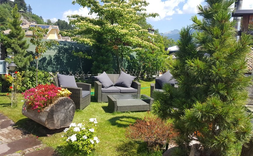 Garten mit Sitzgruppe aus Rattan, bunten Blumen und grünen Bäumen bei Sonnenschein