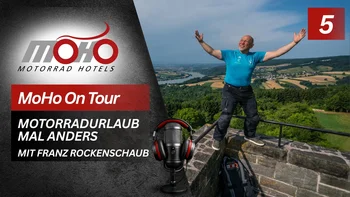 MoHo Podcast Mann in blauer Kleidung auf Aussichtspunkt mit Talblick, Motorradhotel-Logo und Mikrofon