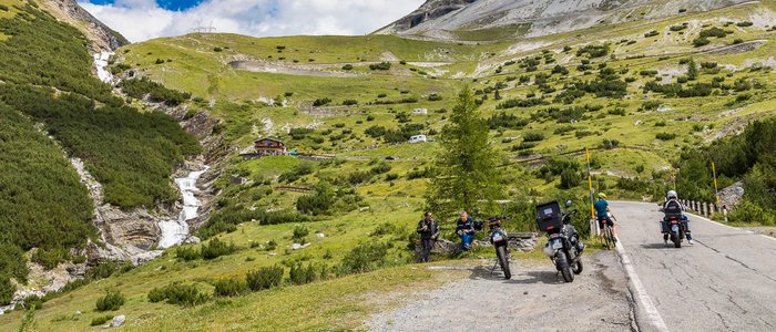 Bergweg met motorrijders, wandelaars en een waterval in de Alpen