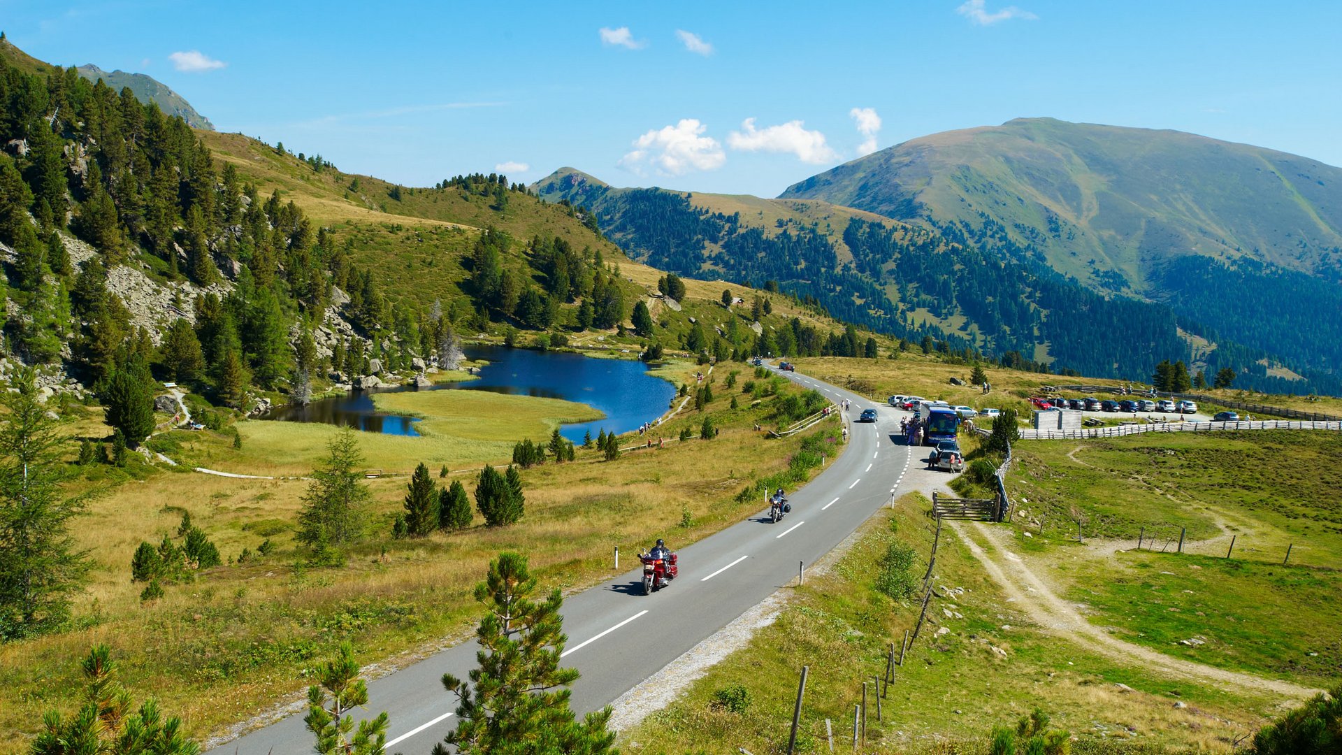 Gebirgsstraße mit Motorradfahrern, See und bewaldeten Bergen im Sommer