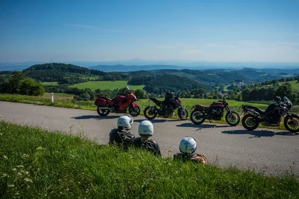 Drie motorrijders zitten aan de weg met uitzicht op groene heuvels en blauwe bergen