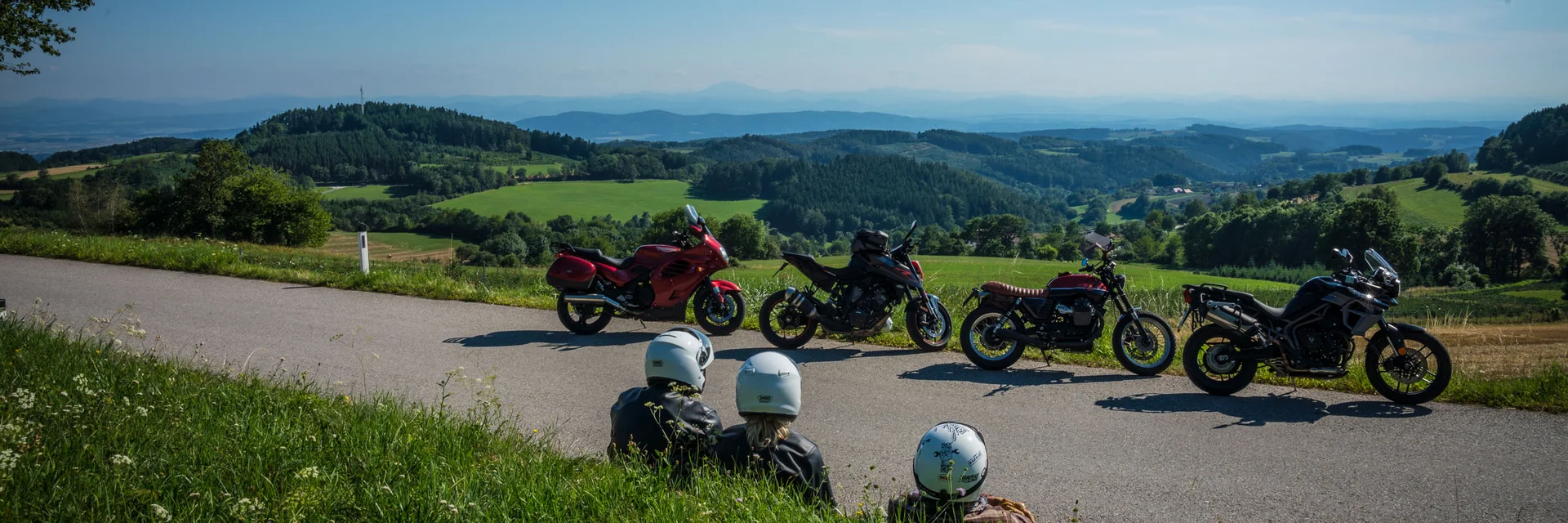 Waldviertel © Moppetfoto.de Drei Motorradfahrer sitzen am Straßenrand mit Blick auf grüne Hügel und blaue Berge