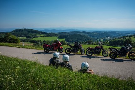 Drei Motorradfahrer sitzen am Straßenrand mit Blick auf grüne Hügel und blaue Berge