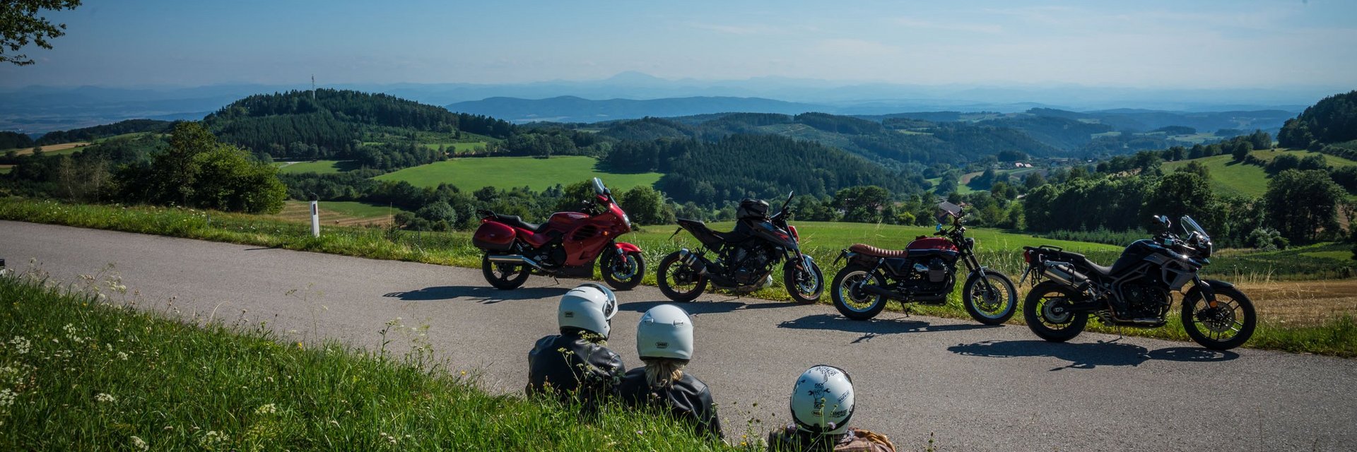 Drei Motorradfahrer sitzen am Straßenrand mit Blick auf grüne Hügel und blaue Berge