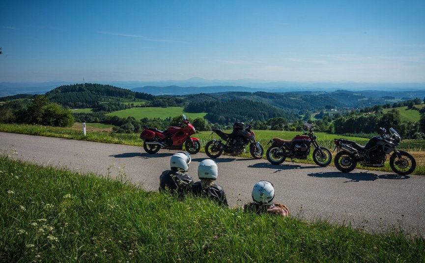 Drei Motorradfahrer sitzen am Straßenrand mit Blick auf grüne Hügel und blaue Berge