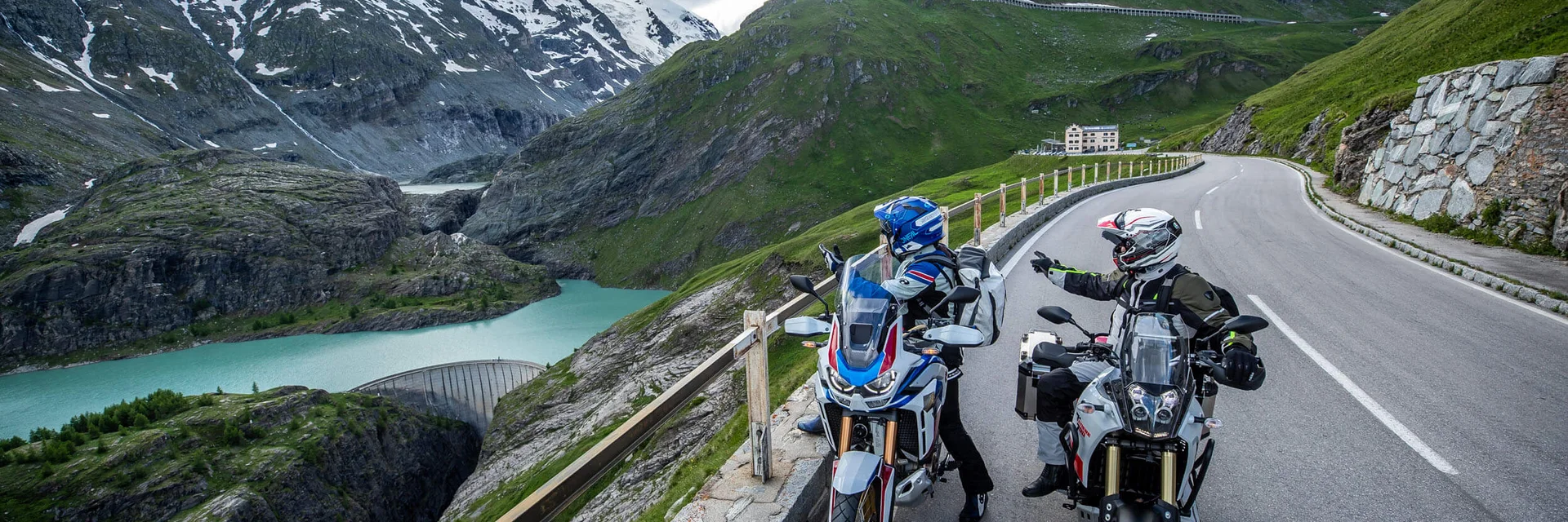 Two motorcyclists on alpine road with mountain lake and snowy peaks