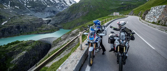 Zwei Motorradfahrer auf Alpenstraße mit Bergsee und schneebedeckten Gipfeln