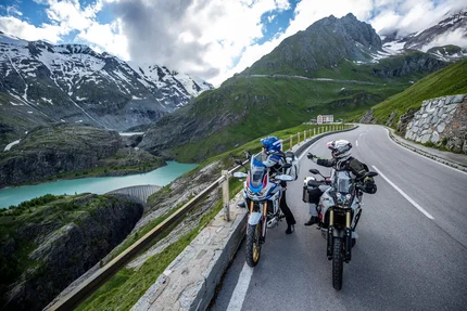Zwei Motorradfahrer auf Alpenstraße mit Bergsee und schneebedeckten Gipfeln